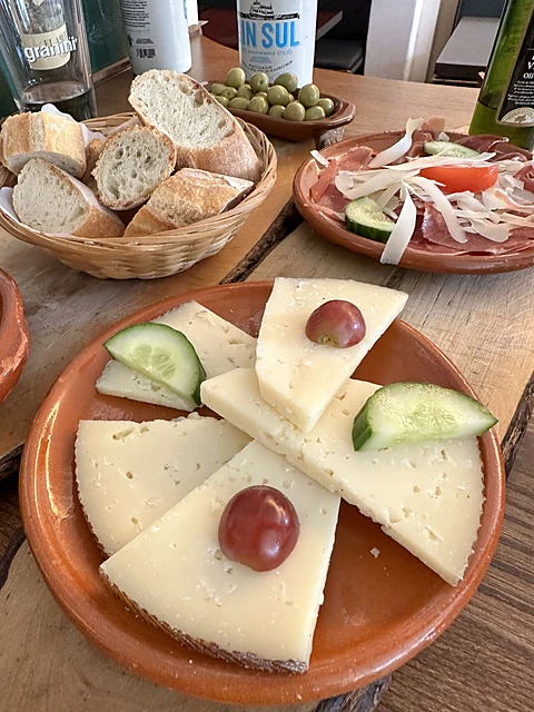Tapas auf Tontellern: Manchego mit Weintrauben und Gurke, Brotkorb, Oliven und Aufschnitt auf einem Holztisch