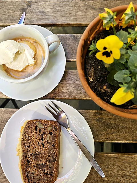 Cappuccino mit Milchschaum und ein Stück Carrot Cake mit Gabel auf einem Holztisch, daneben ein Topf gelber Stiefmütterc