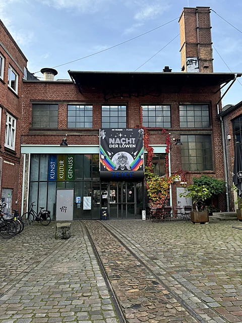 Backsteinfassade einer alten Fabrik mit hohem Schornstein, Glaseingang und alten Schienen im Kopfsteinpflaster