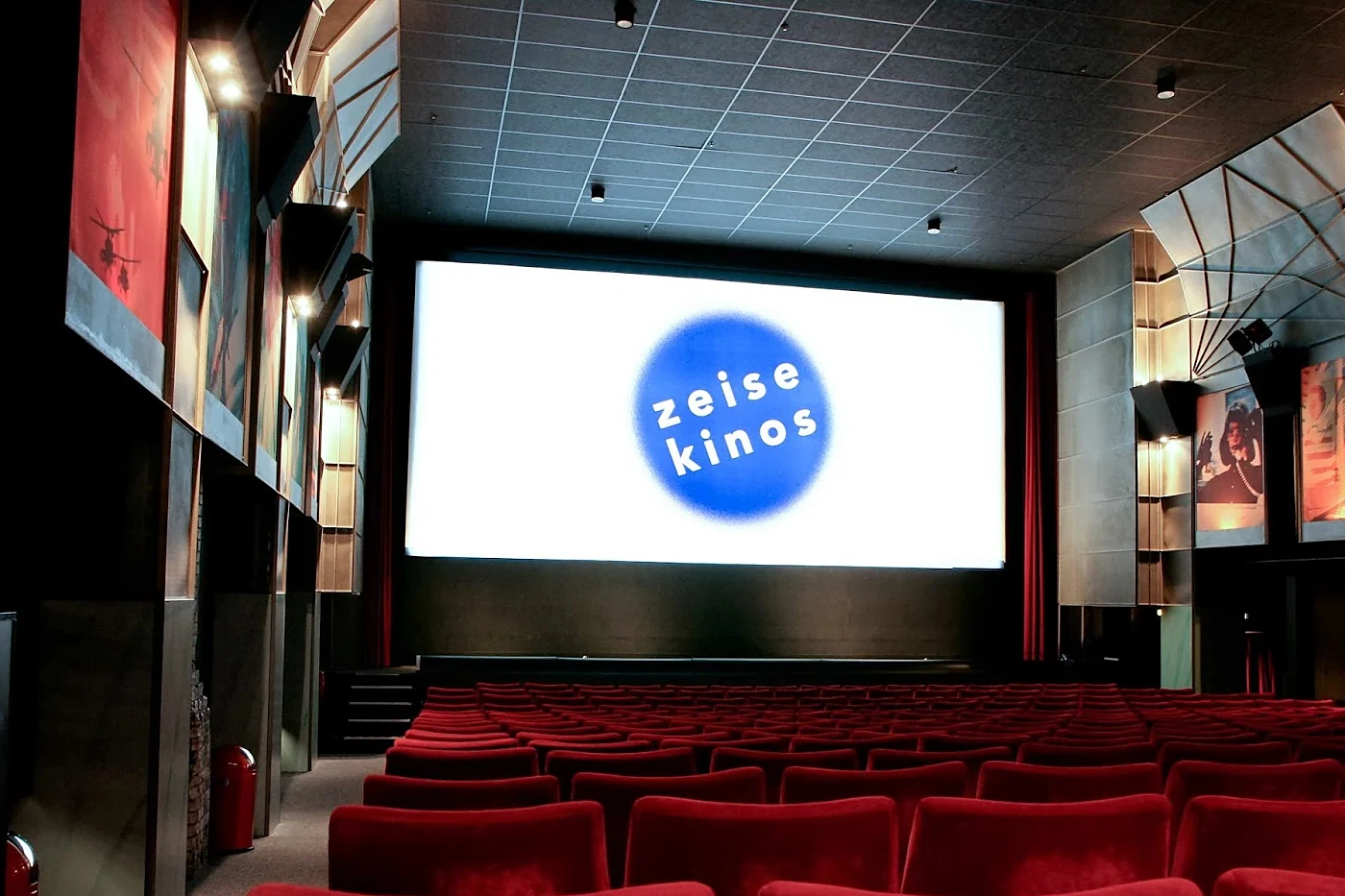 Kinosaal mit roten Sesseln, heller Leinwand mit blauem Zeise-Kinos-Logo und Kunstwerken an den Seitenwänden