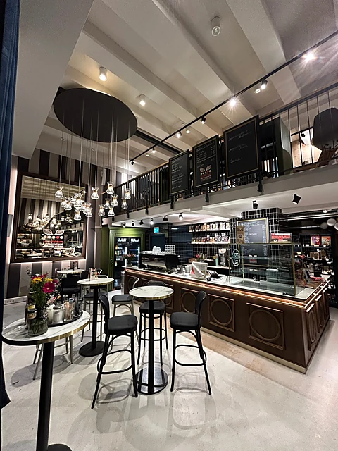 Hohe Café-Halle mit dunkler Holztheke, Kuchenvitrine, Kreidetafeln, Kugellampen und Bistrotischen mit Hockern